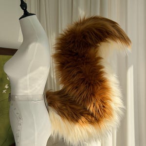 May include: A long, fluffy, faux fur tail in shades of brown and cream, attached to a clear plastic belt. The tail features a thick, voluminous texture with a dark brown base that transitions into a lighter cream color at the tip and underside.