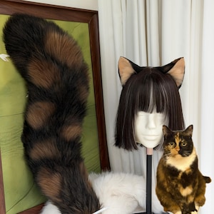 Può includere: Una lunga e soffice coda in pelliccia sintetica con bande alternate nere e marroni, abbinata a una parrucca nera corta e liscia con frangia e orecchie da gatto. Un gatto calico dal pelo nero, arancione e bianco è in primo piano.