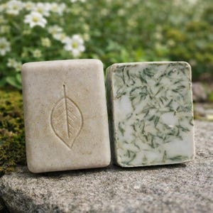 Puede incluir: Dos jabones rectangulares hechos a mano. Uno es de color beige claro con una impresión de hoja. El otro es blanco con inclusiones verdes en forma de hoja. Los jabones se exhiben sobre una superficie de piedra gris, con un fondo borroso de follaje verde y flores blancas.