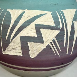 May include: A handcrafted ceramic pot with a turquoise top, a deep purple base, and a cream-colored geometric design. The pot has a rounded shape and a light gray base. The design is carved into the surface.