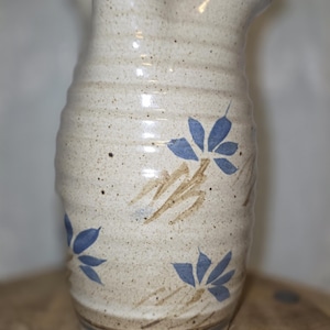 May include: A beige ceramic pitcher with a wavy rim and blue floral designs. The pitcher has a speckled texture and a blue band around the top. The flowers are blue with brown stems.