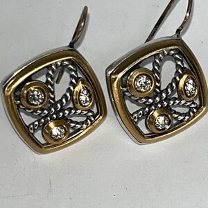 K&ouml;nnte beinhalten: Ein Paar quadratische Ohrringe mit goldfarbenem Rand und silberfarbenem Filigranmuster. Jeder Ohrring hat drei klare, runde Edelsteine und einen goldfarbenen Akzent. Die Ohrringe haben einen Hakenverschluss.