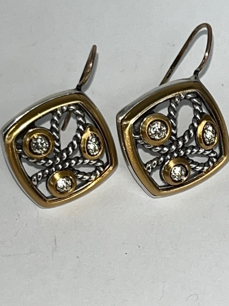 K&ouml;nnte beinhalten: Ein Paar quadratische Ohrringe mit goldfarbenem Rand und silberfarbenem Filigranmuster. Jeder Ohrring hat drei klare, runde Edelsteine und einen goldfarbenen Akzent. Die Ohrringe haben einen Hakenverschluss.