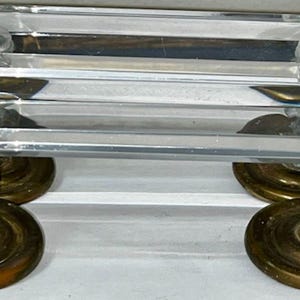 May include: A clear, rectangular crystal object with four brass-colored wheels. The crystal has a layered design with rounded, clear knobs. The wheels are circular and have a vintage appearance. The object appears to be a decorative piece.