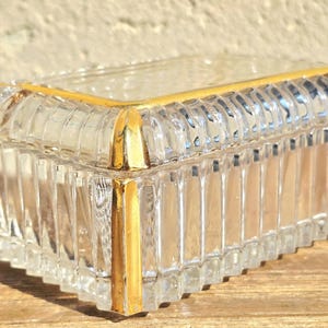 May include: A rectangular clear glass trinket box with a lid, featuring vertical ribbed sides and a gold-colored trim. The box is sitting on a wooden surface, with sunlight casting shadows.