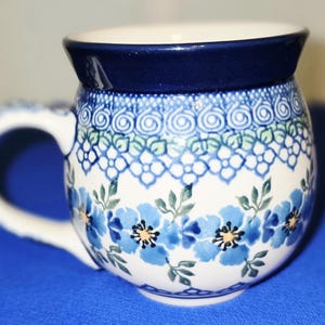 May include: A ceramic mug with a white interior and a dark blue rim. The mug is decorated with blue floral patterns and green leaves. The handle is white. The mug is on a blue surface.