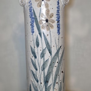 May include: A tall, ceramic vase with a cream-colored base and a blue rim. The vase is decorated with hand-painted flowers and foliage in shades of blue and gray. It features two small handles and measures approximately 12 inches tall.
