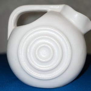 May include: A white ceramic pitcher with a handle and a spout. The pitcher has a spiral design on the side. The pitcher is sitting on a blue surface.