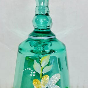 May include: A teal glass bell with a long handle and a flared base. The bell is decorated with a hand-painted floral design featuring a large white and yellow flower with a blue center, and green leaves.