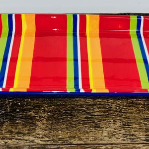 May include: A rectangular serving tray with a vibrant striped pattern in red, orange, yellow, green, and white. The tray has blue handles on each end. The tray is on a dark wooden surface.