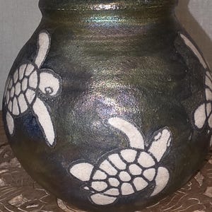 May include: A dark green ceramic vase with white turtle designs. The vase has a round body and a slightly flared rim. The turtles are outlined in black and have detailed shells.