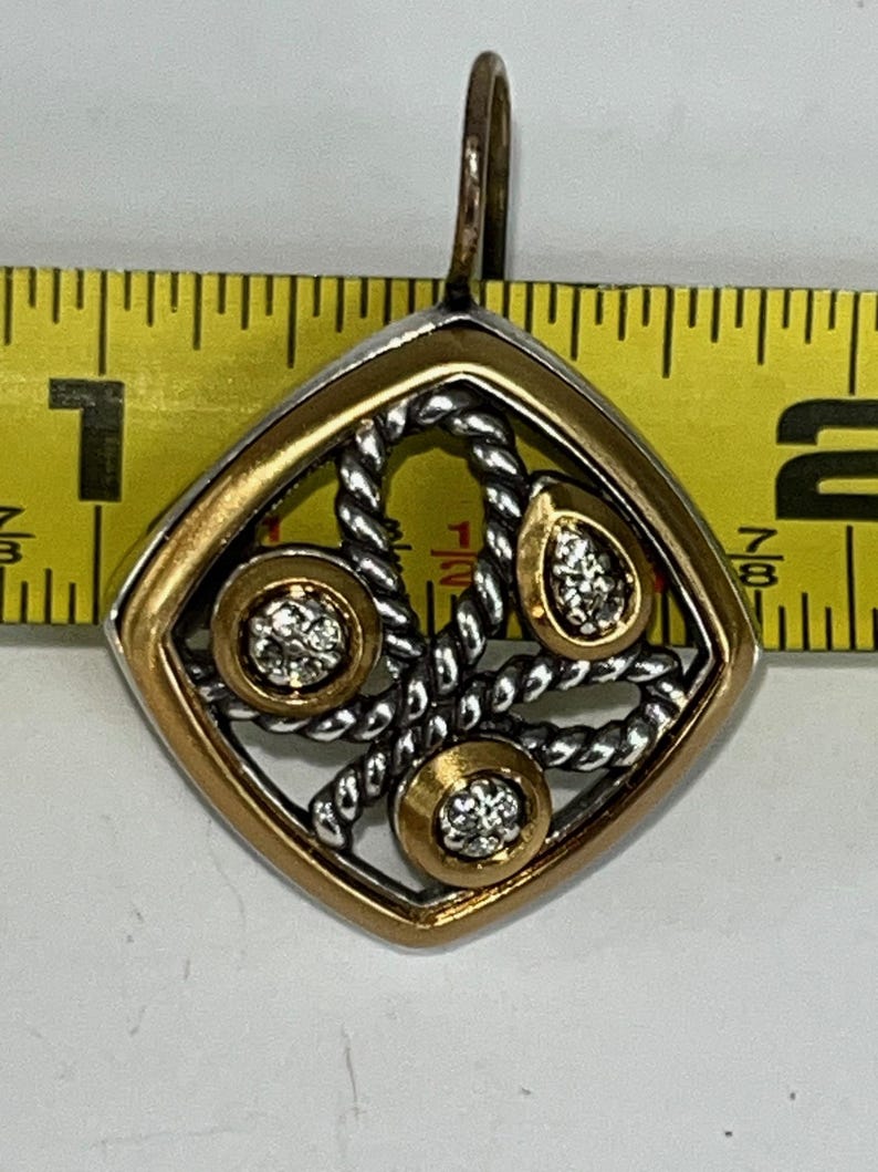 K&ouml;nnte beinhalten: Ein gold-silberner Anh&auml;nger mit einem diamantartigen Edelstein-Design. Der Anh&auml;nger ist quadratisch mit einem goldenen Rand und einem silbernen seilartigen Design in der Mitte. Drei goldene Kreise mit Edelsteinen sind in das Design eingesetzt. Der Anh&auml;nger ist etwa 2,5 cm lang.