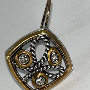 K&ouml;nnte beinhalten: Einzelner Ohrring mit Gold- und Silberdesign. Der Ohrring hat einen quadratischen Rahmen mit goldenem Rand, der ein silbernes, seilartiges Muster umschlie&szlig;t. Drei goldgerahmte Fassungen halten klare Edelsteine.