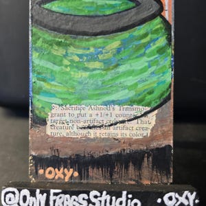 May include: Hand-painted artwork of a Magic: The Gathering card, "Ashnod's Transmogrant." The card features a green and black cauldron design with text. The artist's signature "OXY" is visible, along with "@Only FRAGS Studio."