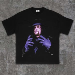 Peut inclure: T-shirt noir avec un graphisme d'un homme portant un chapeau et des gants violets. L'image est un portrait d'un lutteur aux longs cheveux noirs et à l'expression sévère. Le t-shirt a un col rond et des manches courtes.