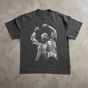 May include: Black t-shirt featuring a black and white image of a bald man with his arms raised. The man is wearing a vest with a skull and the letters "BMF". The shirt is made of a soft material.