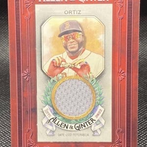 Op de afbeelding: Een 2022 Topps Allen & Ginter honkbalkaart met een portret van David Ortiz. De kaart heeft een rode rand met de tekst "ORTIZ" en "GAME-USED MEMORABILIA". De kaart heeft ook een stoffen patch.