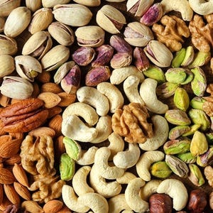May include: A variety of mixed nuts including shelled pistachios, cashews, walnuts, pecans, and hazelnuts. The assortment features a mix of beige, brown, and green colors with varied textures, showcasing whole nuts and kernels arranged in a dense, overlapping pattern.