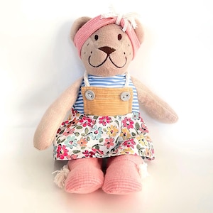 May include: A plush teddy bear wearing a pink headband, a blue and white striped top, and a floral print skirt. The bear has pink socks and a corduroy apron. The bear is a soft toy.