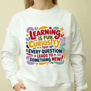 May include: White sweatshirt with a colorful graphic that reads "LEARNING is FUN, CURIOSITY too! EVERY QUESTION + LEADS TO + SOMETHING NEW!" The design features red, blue, yellow, green, and purple text and shapes.