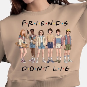 Può includere: Felpa beige con la scritta "FRIENDS DON'T LIE" e una grafica di sei personaggi. I personaggi indossano vari outfit, tra cui giacche, pantaloncini e magliette. La felpa ha un girocollo a coste.
