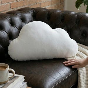 Puede incluir: Un cojín decorativo blanco con forma de nube descansa sobre un sofá de cuero marrón oscuro. La almohada es esponjosa y de aspecto suave. Una pila de libros y una taza de café están sobre una pequeña mesa auxiliar de madera. Una mano toca una manta color crema.