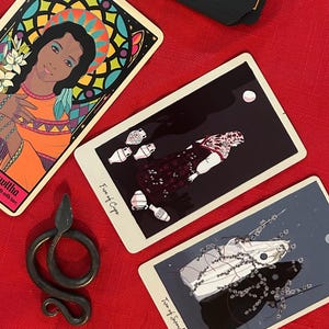 May include: An overhead shot of tarot cards and a decorative metal snake on a red surface. The cards feature colorful illustrations and text, including "Five of Cups" and "Two of Swords". A deck of cards with a floral design and a deck with a stained glass design are also visible.