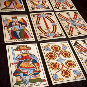 Puede incluir: Un conjunto de cartas de tarot con ilustraciones coloridas y texto en francés. Las cartas muestran figuras y símbolos en una paleta de rojo, azul, amarillo y beige. Las cartas están dispuestas sobre una superficie oscura. El texto incluye "IVSTICE".