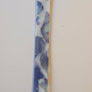 May include: A rectangular, handmade resin bookmark with a hole at the top. The bookmark features a blend of blue, white, and iridescent glitter, creating a marbled effect. The design is abstract and fluid, with a glossy finish.