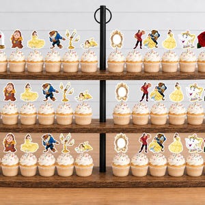 May include: A three-tiered wooden cupcake stand holding rows of cupcakes topped with paper cutouts featuring characters and objects from Beauty and the Beast, including a red rose, a mirror, a teapot, and figures of Belle and the Beast.