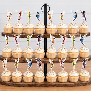 May include: A three-tiered dark wood cupcake stand holding twenty-four cupcakes, each topped with a colorful fairy-shaped paper topper. The cupcakes feature white frosting with multi-colored sprinkles and are arranged in rows on the wooden tiers.