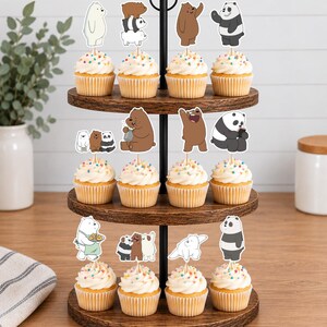 May include: A three-tiered wooden cupcake stand holding twelve cupcakes topped with white frosting and colourful sprinkles. Each cupcake features a paper topper depicting cartoon bears in various poses, including stacked bears and individual characters.