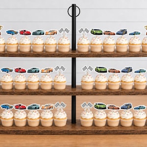 May include: A three-tiered wooden stand holding cupcakes decorated with paper toppers featuring various colorful race cars, checkered flags, and gold trophies. The cupcakes are arranged in rows on each level of the dark brown tiered display stand.