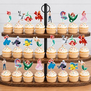 Puede incluir: Un soporte de madera para cupcakes de tres niveles con veinticuatro cupcakes, cada uno decorado con un recorte de papel colorido de personajes de La Sirenita, incluyendo a Ariel, Flounder, Sebasti&aacute;n, Trit&oacute;n, &Uacute;rsula y Eric.