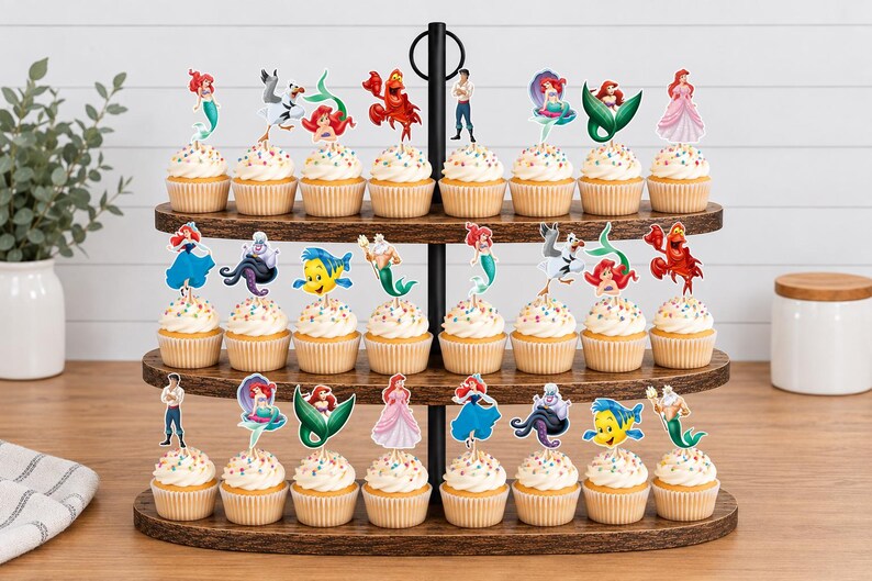 Puede incluir: Un soporte de madera para cupcakes de tres niveles con veinticuatro cupcakes, cada uno decorado con un recorte de papel colorido de personajes de La Sirenita, incluyendo a Ariel, Flounder, Sebasti&aacute;n, Trit&oacute;n, &Uacute;rsula y Eric.