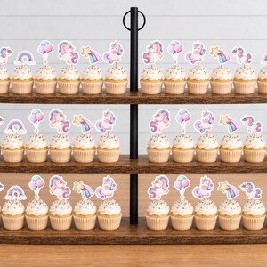 May include: A three-tiered wooden cupcake stand holding numerous cupcakes topped with decorative paper toppers featuring pastel-coloured illustrations of unicorns, rainbows, stars, and balloons. The stand has a black metal central handle.