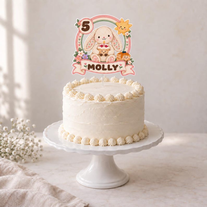 May include: A white frosted cake topped with a decorative topper featuring a cartoon bunny holding a cupcake, a sun, a rainbow, the number 5, and a banner with the name MOLLY printed in brown text, all accented with small colorful flowers.