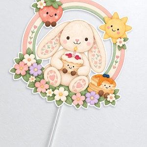 K&ouml;nnte beinhalten: Ein Tortenstecker mit einem cremefarbenen Hasen, der einen kleinen Kuchen mit Kerze h&auml;lt, umgeben von einem pastellfarbenen Regenbogen, einer l&auml;chelnden Erdbeere, einer gelben Sonne und bunten Blumen, befestigt an einem transparenten Kunststoffstab.