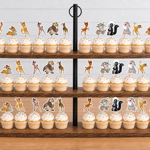 May include: A three-tiered wooden cupcake stand holding numerous cupcakes topped with decorative paper cutouts of forest animals, including a fawn, a rabbit, and a skunk. The stand features a central black metal rod with a circular handle at the top.
