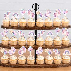 May include: A three-tiered wooden cupcake stand holding twenty-two cupcakes, each topped with a decorative paper cutout featuring watercolour illustrations of unicorns, rainbows, shooting stars, and pastel balloons on a white background.