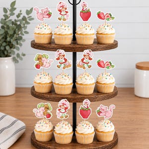 Op de afbeelding: Een houten cupcake-standaard met drie niveaus en twaalf cupcakes met witte glazuur en kleurrijke hagelslag. Elke cupcake is voorzien van een papieren topper met een stripfiguur in een rode hoed, roze katten en aardbeien.