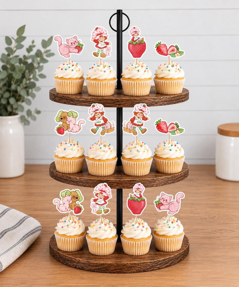 Op de afbeelding: Een houten cupcake-standaard met drie niveaus en twaalf cupcakes met witte glazuur en kleurrijke hagelslag. Elke cupcake is voorzien van een papieren topper met een stripfiguur in een rode hoed, roze katten en aardbeien.