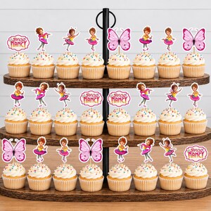 May include: A three-tiered wooden stand holding twenty-four cupcakes topped with Fancy Nancy themed paper decorations. The toppers feature illustrations of a girl in a pink outfit, pink butterflies, and the Fancy Nancy logo in pink and white.