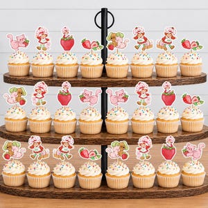 Op de afbeelding: Een houten standaard met drie niveaus en vierentwintig cupcakes, elk voorzien van een papieren uitsparing met Strawberry Shortcake-figuren, roze katten en aardbeien. De cupcakes hebben witte glazuur met kleurrijke spikkels in papieren vormpjes.