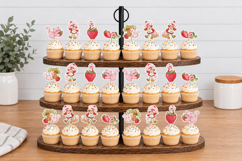 Op de afbeelding: Een houten standaard met drie niveaus en vierentwintig cupcakes, elk voorzien van een papieren uitsparing met Strawberry Shortcake-figuren, roze katten en aardbeien. De cupcakes hebben witte glazuur met kleurrijke spikkels in papieren vormpjes.