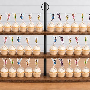 May include: A three-tiered wooden cupcake stand holding numerous cupcakes topped with colorful fairy-shaped paper decorations. The stand features a central black metal pole with a circular handle at the top, displaying rows of treats with various fairy figures.
