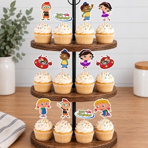 Puede incluir: Un soporte de madera de tres niveles con doce cupcakes cubiertos de glaseado blanco y chispas de colores. Cada cupcake tiene un adorno de papel de Little Einsteins con personajes como Leo, June, Quincy, Annie y la nave espacial roja.