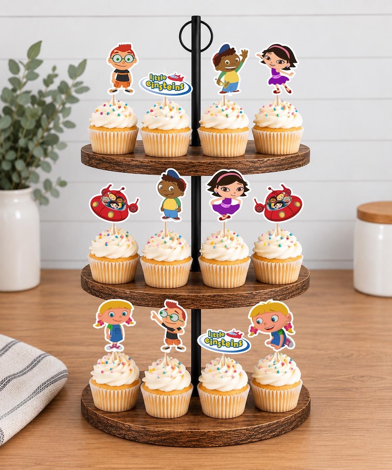 Puede incluir: Un soporte de madera de tres niveles con doce cupcakes cubiertos de glaseado blanco y chispas de colores. Cada cupcake tiene un adorno de papel de Little Einsteins con personajes como Leo, June, Quincy, Annie y la nave espacial roja.