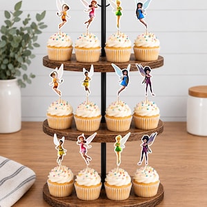 May include: A three-tiered wooden cupcake stand holding twelve cupcakes, each topped with a decorative fairy cutout. The cupcakes feature white frosting with colorful sprinkles. The stand has a black metal central pole and circular wooden shelves.