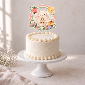 K&ouml;nnte beinhalten: Ein wei&szlig; glasierter Kuchen mit einem Tortenstecker, der einen Cartoon-Hasen mit einem Cupcake zeigt, umgeben von einem Regenbogen, einer l&auml;chelnden Erdbeere, einer Sonne und bunten Blumen auf einem wei&szlig;en Tortenst&auml;nder.